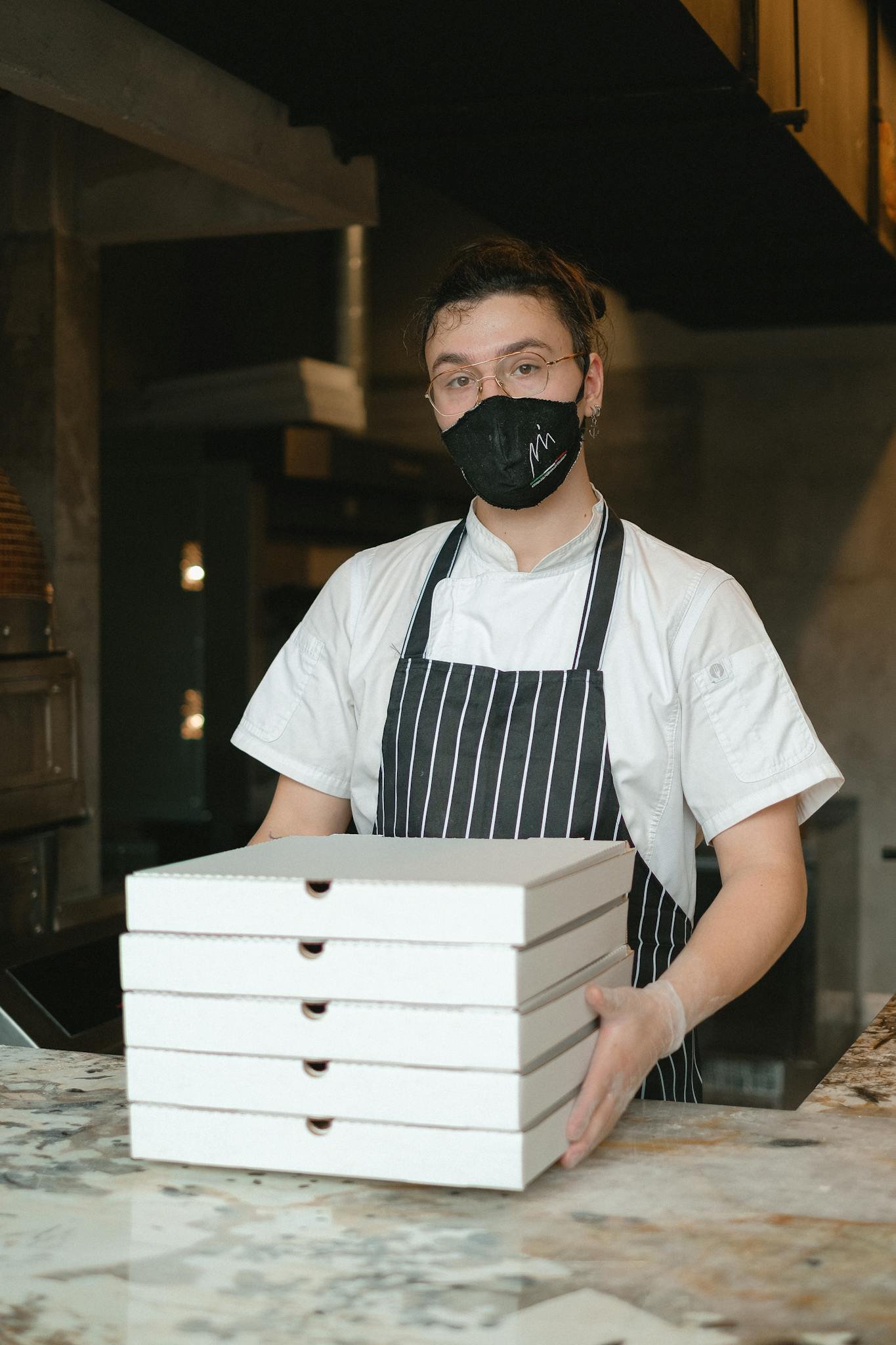 A chef in a restaurant kitchen holding a stack of pizza boxes while wearing a face mask.