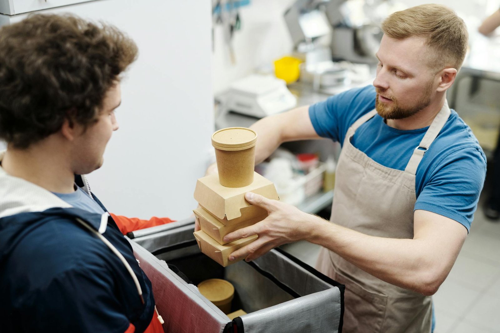 A delivery worker in an apron packs food containers into a bag in a kitchen setting.