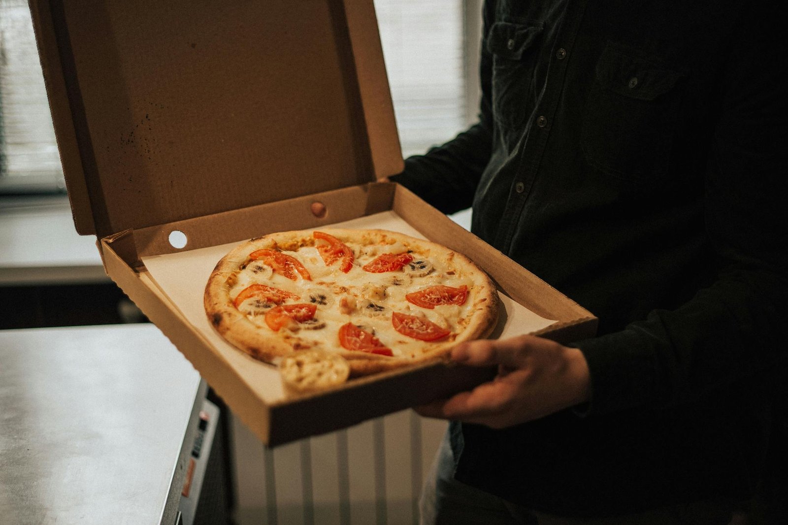 A person holds an open box with a freshly baked pizza topped with tomatoes and cheese.
