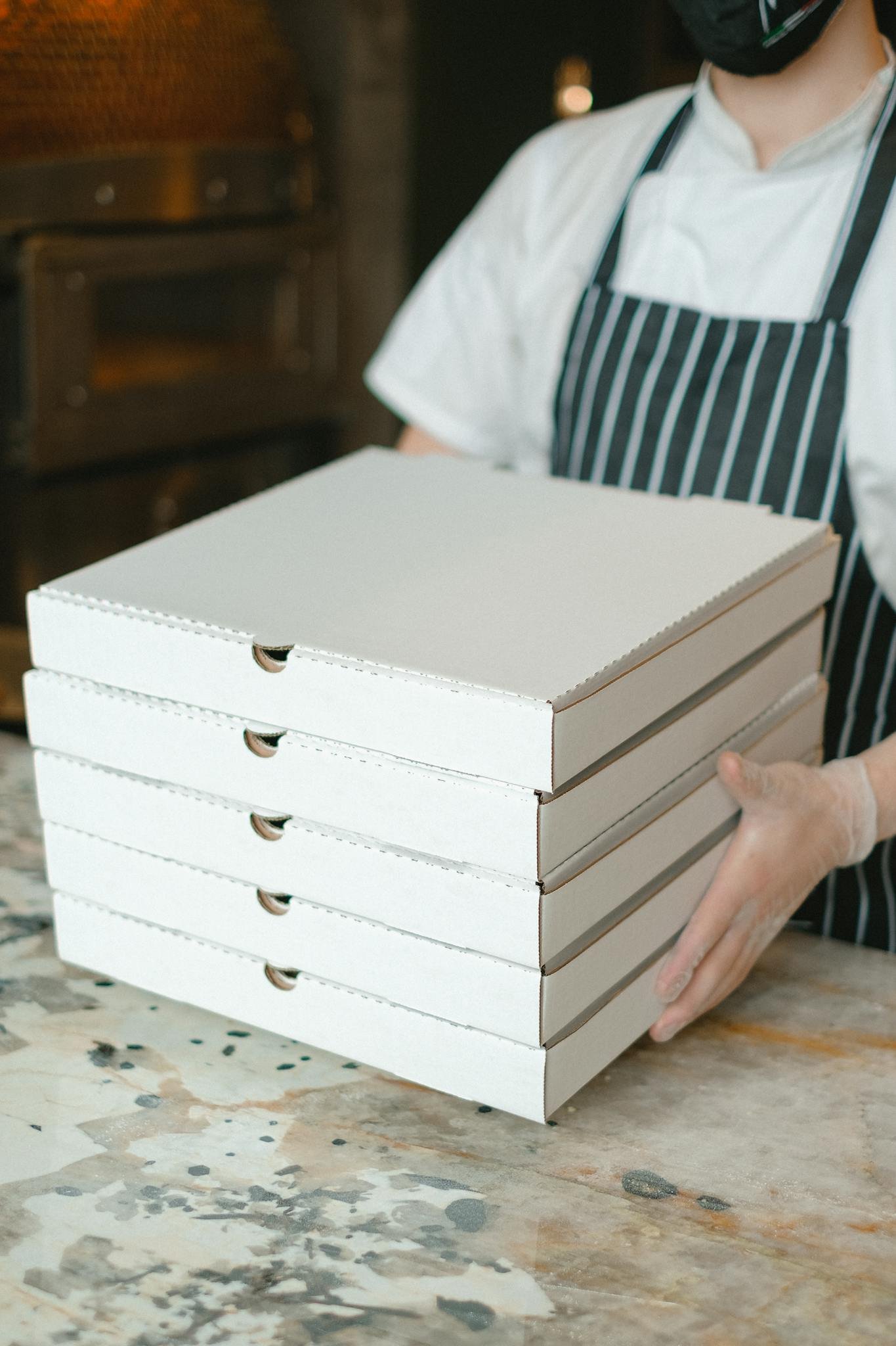 A pizza delivery worker holds stacked boxes in a restaurant kitchen, ready for delivery.