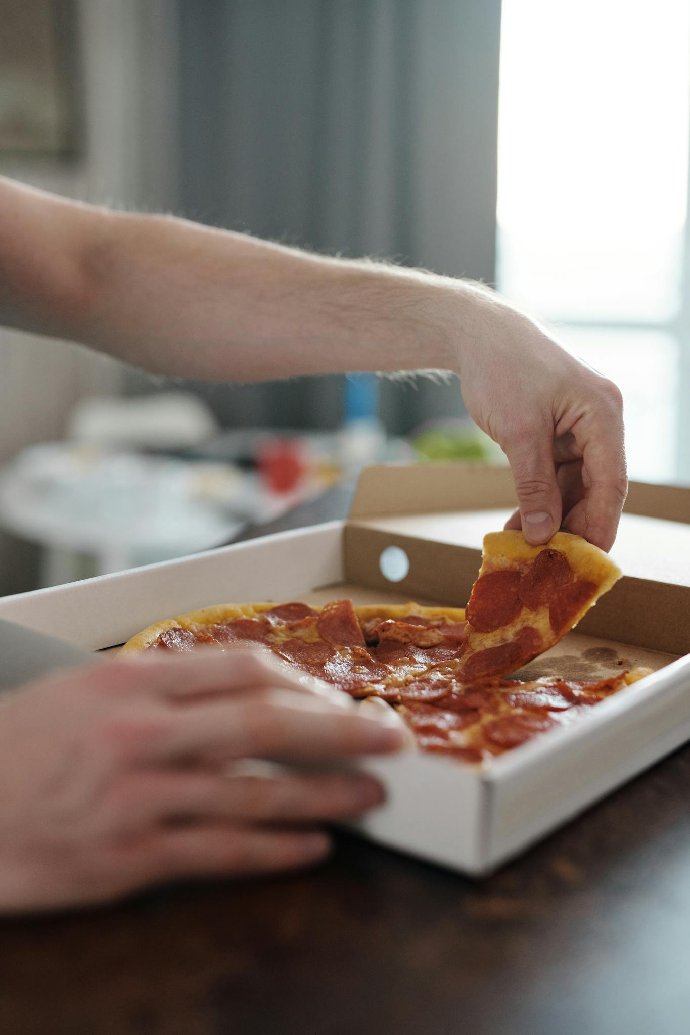 Close-up of hands picking a slice of pepperoni pizza from a box, showcasing a delicious and casual dining moment.
