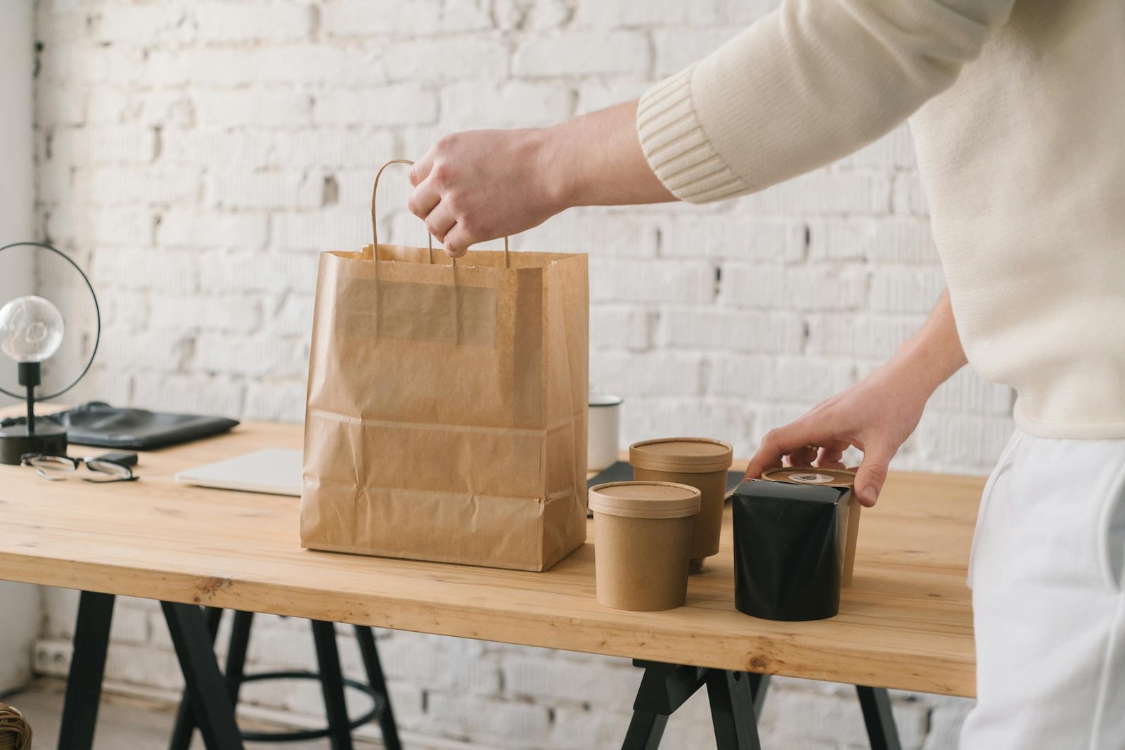 Hand arranging takeaway food packaging on a wooden table with brick wall background.